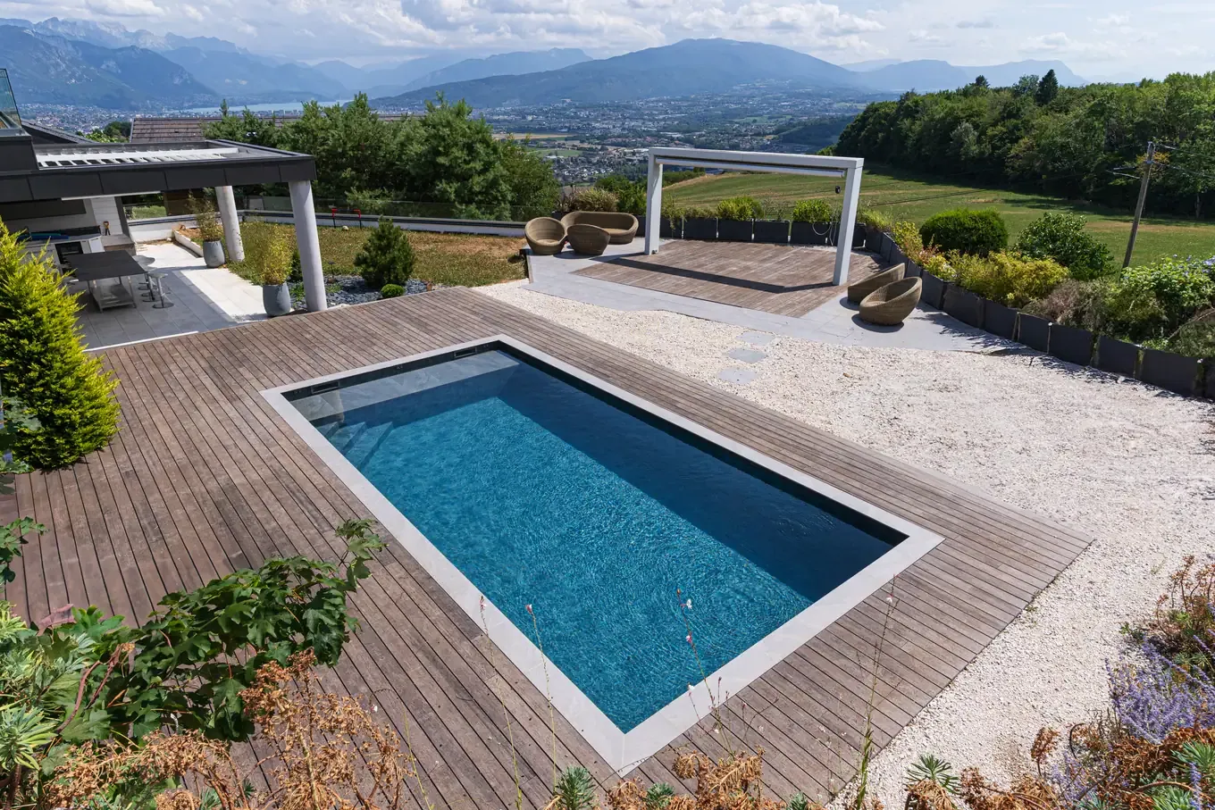 Piscine sur mesure bordée d’une terrasse en bois et d’une pergola, face aux montagnes, à Metz-Tessy