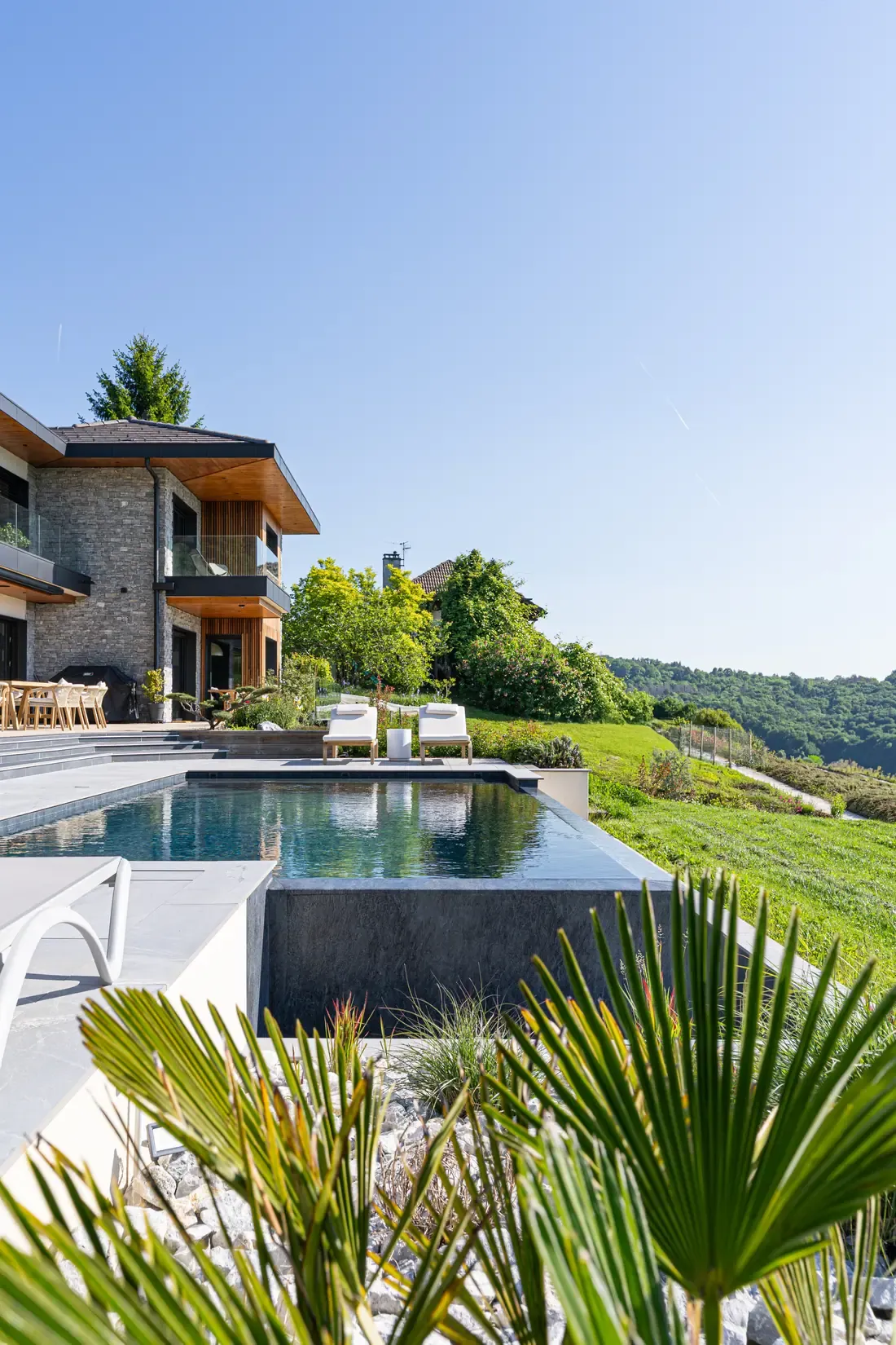 Piscine à débordement devant une maison contemporaine en pierre et bois, avec transats face à la vallée, à Pringy