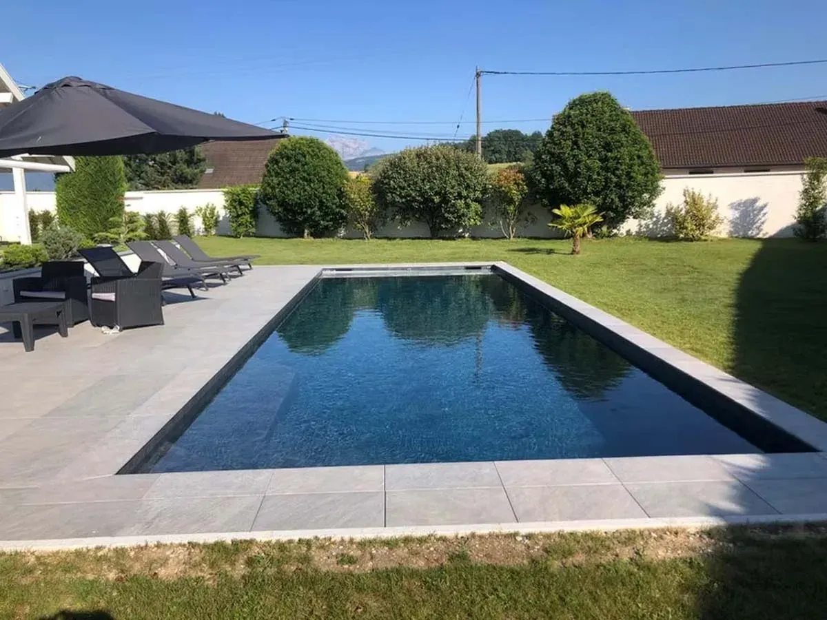 Piscine rectangulaire à revêtement gris foncé, entourée d’une terrasse en pierre avec transats, sous un ciel bleu