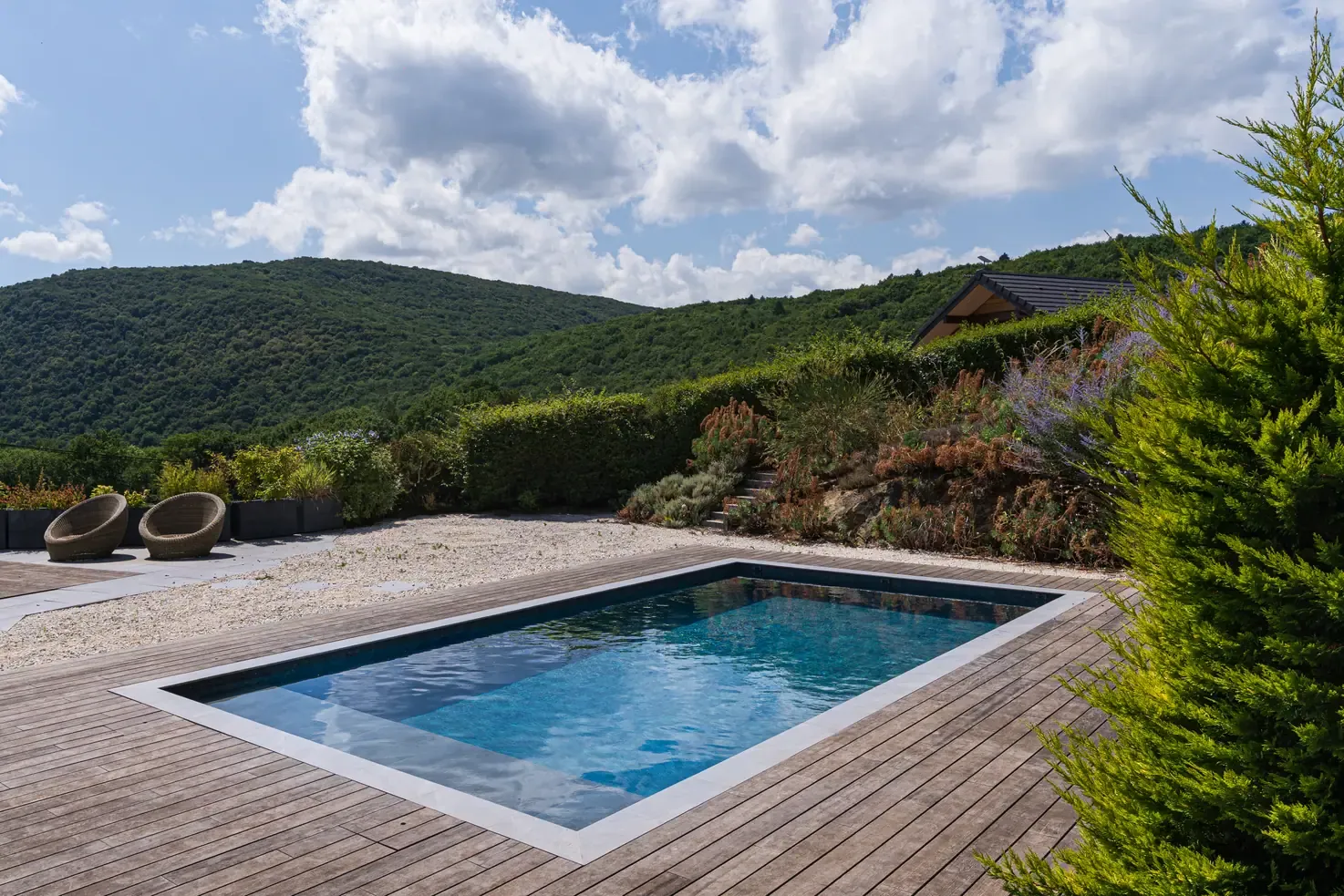 Piscine avec margelles claires et terrasse en bois devant un jardin arboré à Metz-Tessy
